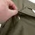 Nahaufnahme einer Hand, die eine grüne Stofflasche mit einem Schlitz anhebt. Detailansicht des Materials und der Verarbeitung.