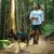 Mann führt einen Hund an der Leine durch einen Waldweg. Der Hund trägt ein Geschirr.