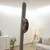 Chat accroché à un grand poteau à griffer gris, fixé du sol au plafond dans un salon moderne avec canapé, lampe et plante. Le matériau du poteau semble être en tissu.