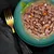Close-up of a green plate with pieces of meat, diced carrots and white vegetable cubes in sauce, garnished with basil. Golden fork and black cloth beside it.