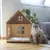 Wooden house with window design and padded lying surface, suitable as pet accommodation. Cat sitting next to it on wooden floor.