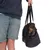 Petit chien à poil court dans un sac de transport noir porté à la main par une personne en short vert et sweat bleu. Sac avec anses réglables, ouverture pour la tête du chien.