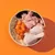 Bowl containing raw chicken drumstick, diced raw meat, sliced carrots, rice and dark leafy pieces, showing visible ingredients of pet food.