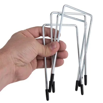 Mano sujetando varias estacas metálicas en forma de U con extremos recubiertos de plástico negro, visibles cinco unidades.