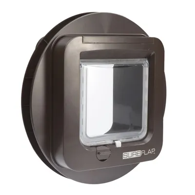 Round, dark-coloured pet door with clear flap and locking mechanism, shown from the front. The word 'SureFlap' is visible on the bottom right corner.