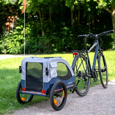 Przyczepka rowerowa dla zwierząt z oknami z siatki, niebieska i szara tkanina, przymocowana do roweru. Odpowiednia do transportu na ścieżkach lub w parkach.