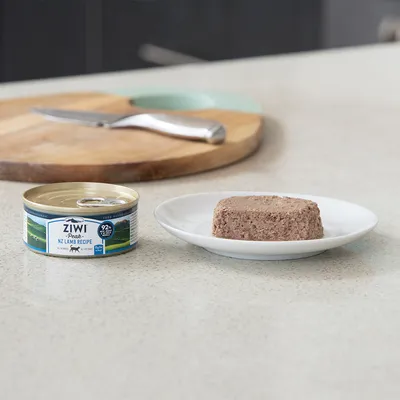 Boîte de pâtée Ziwi Peak NZ Lamb Recipe 92 % viande visible, à côté d'une portion servie sur une assiette blanche. Texte anglais sur l'emballage.