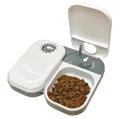 Automatisk foderdispenser med to rum, et åbent låg og en timer med tal fra 0 til 48. Skål med tørfoder synlig i det ene rum. Hvidt og gråt plastmateriale.