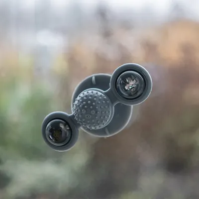 Šedá plastová hračka s přísavkou na okně, dvě otočné části a střed s gumovými výstupky. Viditelné detaily materiálu a konstrukce.