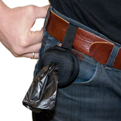 Black bag dispenser with belt clip, attached to a belt. Black waste bags are pulled from the opening.