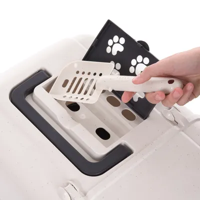 Hand hält eine Streuschaufel, die in ein seitliches Fach einer Katzentoilette mit Pfotenmuster eingesetzt wird.
