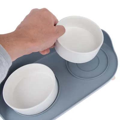Mão levanta uma tigela de comida branca de um suporte cinzento com dois lugares para tigelas.
