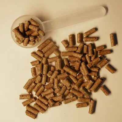 Rjavi peleti za hišne ljubljenčke, razsuti na površini in v prozorni merilni žlički z oznako 5 ml. Vidna je tekstura in oblika hrane.