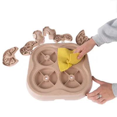 Hand torkar av en beige aktivitetsleksak i plast med gul trasa, flera lösa delar synliga runtom. Leksaken har fyra runda fack.