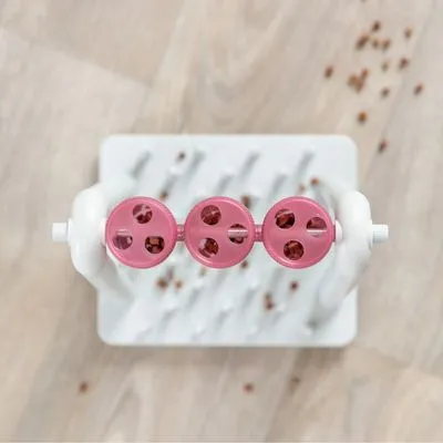 Interactief voerspeelgoed van wit plastic met drie roze draaibare elementen, gevuld met brokjes. Basis met opstaande pinnen zichtbaar. Brokjes verspreid op houten vloer.