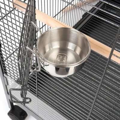 Gamelle en métal fixée à la porte d'une cage grillagée, perchoir en bois visible à l'intérieur.