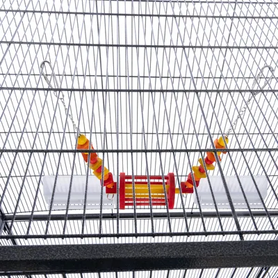 Colivie metalică cu jucărie suspendată din plastic roșu și galben, agățată pe lanțuri metalice. Jucăria are formă cilindrică și elemente rotunde colorate.