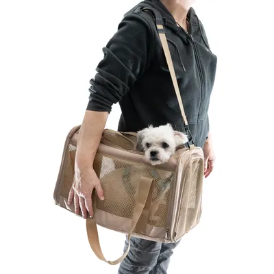 Sac de transport beige à mailles, porté en bandoulière par une personne, avec un petit chien blanc visible à l’intérieur. Poignées et sangle réglable apparentes.