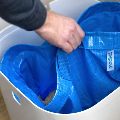 Blauwe kunststof binnenzak met handvat en zichtbaar merklabel 'modkat' in witte kattenbak, hand houdt de zak vast.