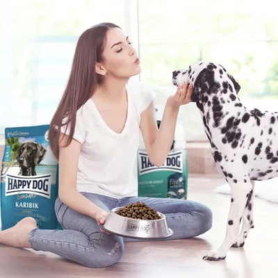 Happy Dog Karibik torrfoder i skål, dalmatiner matas. Förpackning i bakgrunden visar varumärke och produktnamn.