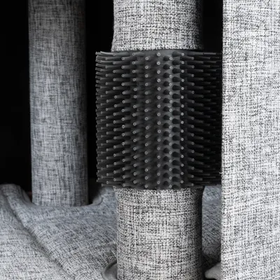 Brosse noire en plastique fixée autour d’un poteau recouvert de tissu gris, partie d’un arbre à chat ou griffoir visible sur l’image.