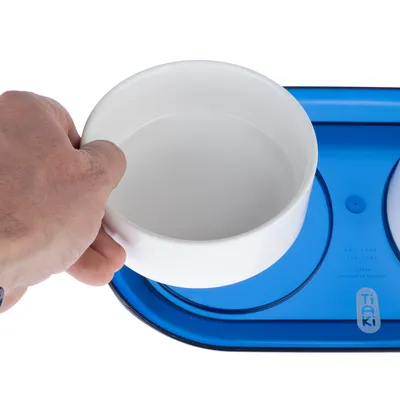 Hand hält weissen Napf über blauem Tiaki-Napfhalter mit zwei runden Aussparungen.
