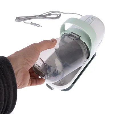 Hand holding transparent container of an electric device with cable and green handle. Device shows on/off button.