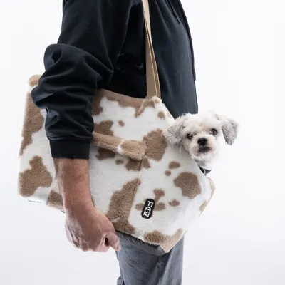 Sac de transport en tissu à motif tacheté beige et blanc, marque TIKI, porté à l’épaule par une personne, petit chien blanc à poils bouclés visible dans le sac.