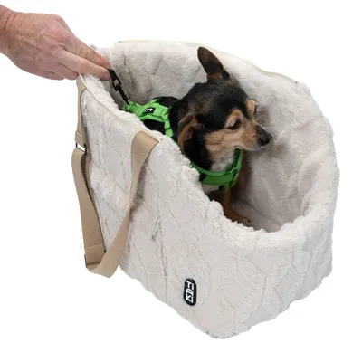 Petit chien avec harnais vert assis dans un sac de transport en tissu beige à bandoulière, marque Trixie visible sur le côté. Main tenant une attache intérieure du sac.