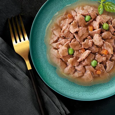 Chunks of meat in gravy with peas and carrot pieces served on a green plate, showing the texture and ingredients of the pet food.