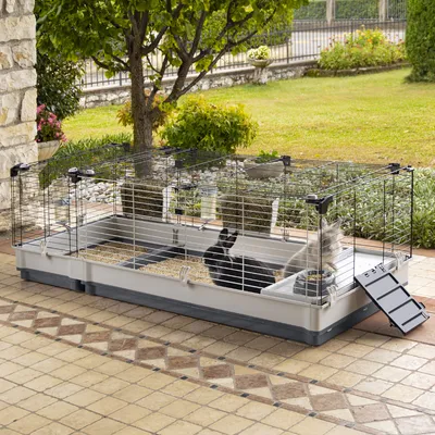 Grosser Käfig mit Metallgitter und Kunststoffboden, geeignet für Kleintiere. Enthält zwei Futterbereiche, Einstreu und eine Rampe. Zwei Tiere sichtbar im Käfig.