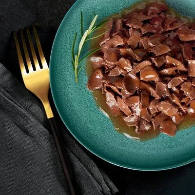 Bord met natvoer in stukjes en jus, geserveerd op een groen bord met een takje rozemarijn. Gouden vork en zwarte servet zichtbaar naast het bord.