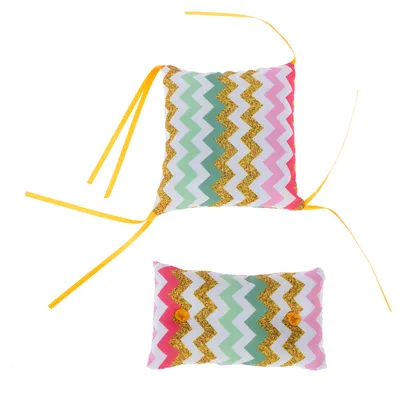 Dos cojines de juego con estampado de zigzag en verde, rosa, blanco y dorado. Cintas amarillas en un cojín, pompones amarillos en el otro.