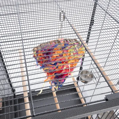 Jouet suspendu en carton et papier multicolore dans une cage à oiseaux en métal avec perchoirs en bois et gamelle en inox visibles.