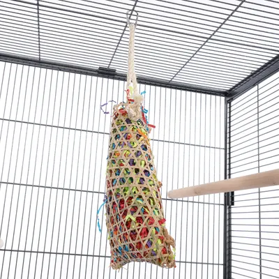 Jouet suspendu en corde et fibres naturelles tressées, rempli de bandes colorées, accroché dans une cage à oiseaux avec perchoir en bois visible.