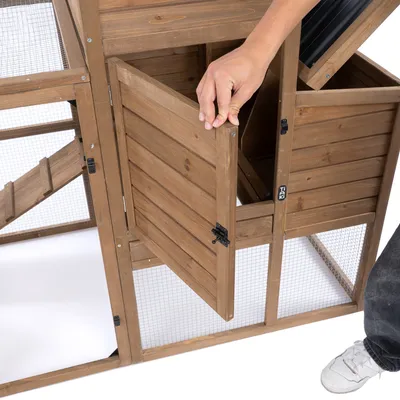Drevený výbeh s mrežovanými stenami, otvorené dvere a viditeľný uzamykací mechanizmus. Ruka otvára dvere, ukazuje jednoduché ovládanie.