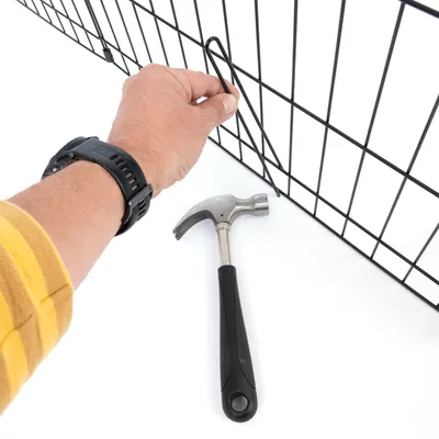 Hand befestigt Metallhaken an einem Gitter mit Hammer auf weissem Hintergrund.