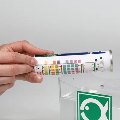 Dłoń trzyma tubę z paskami testowymi i skalą kolorów do analizy wody nad akwarium. Zielony symbol ryby widoczny na akwarium.