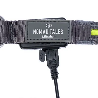 Collier en nylon gris avec étiquette Nomad Tales München, port USB visible pour recharge, détail d’un câble branché.