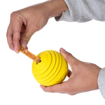 Balle jaune en plastique tenue à deux mains, une main insère une friandise allongée dans l’ouverture de la balle. Motif d’os visible sur le côté de la balle.