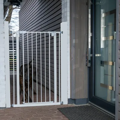Barrière de sécurité blanche en métal installée à l’extérieur devant une porte, chien visible derrière la barrière.