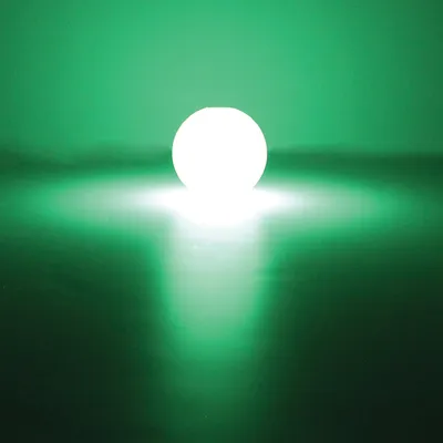 Balle lumineuse ronde émettant une forte lumière blanche avec halo vert visible autour, posée sur une surface sombre. Aucun texte ou marque visible.