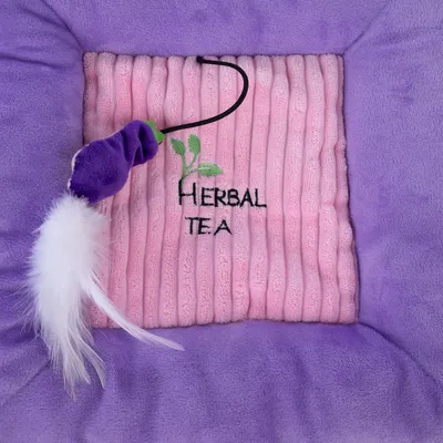 Katzenspielzeug mit lila Stoff, weisser Feder und Stickerei 'Herbal Tea' auf rosa Plüschunterlage.