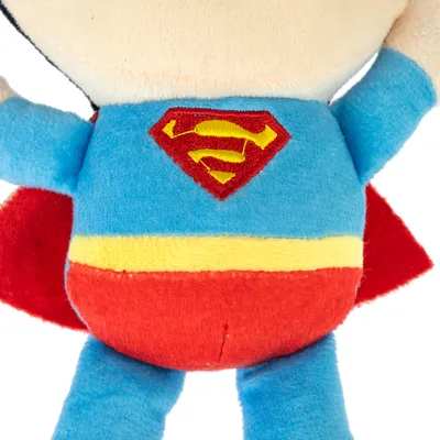 Plyslegetøj i Superman-design med blå dragt, rød kappe og gul bælte. Rødt Superman-logo synligt på bagsiden.