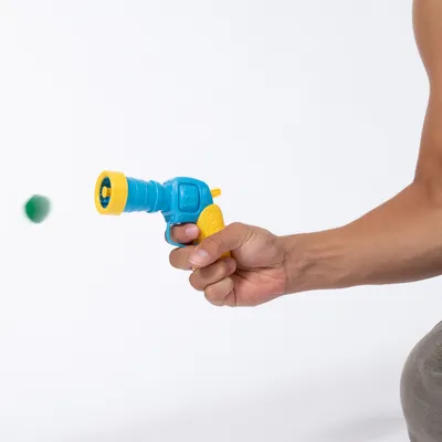 Hand hält eine blaue und gelbe Spielzeugpistole, die einen grünen Ball abschießt.
