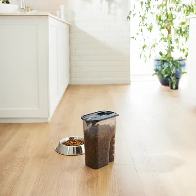 Contenedor de comida transparente con tapa negra, lleno de pienso. Al lado, un cuenco plateado con pienso sobre suelo de madera en una cocina.