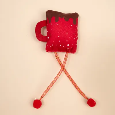 Red fabric toy in the shape of a cup with a brown top, white dots, and two yellow-red ribbons with red pom-poms.