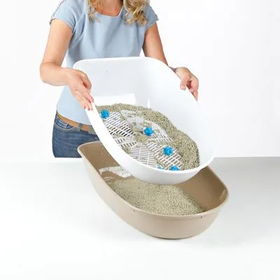 Kattenbak met zeefinzet, vrouw giet kattengrit van witte bak met rooster en blauwe elementen in beige onderbak. Beide bakken van kunststof, grit zichtbaar.