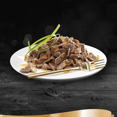 Prato branco com pedaços de carne em molho gelatinoso, decorado com tiras verdes e acompanhado por garfo e faca dourados. Não há marcas ou texto visíveis.