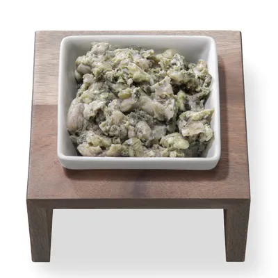 Weiße Keramikschale mit Nassfutter auf einem Holzständer. Sichtbare Zutaten: Fleischstücke und grüne Gemüsestücke.